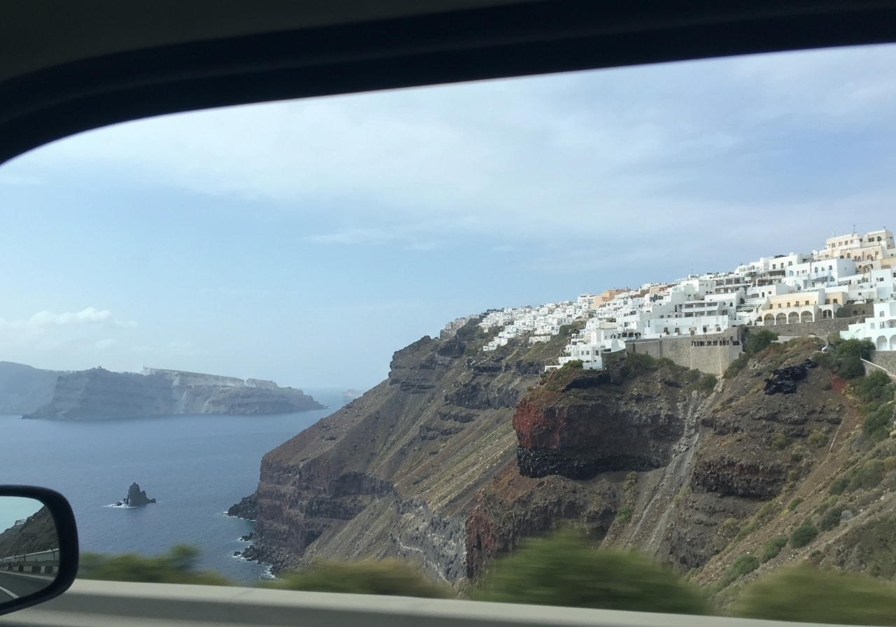 A handheld smartphone shot capturing an authentic moment from a moving taxi. The focus is slightly soft on a section of the dramatic volcanic cliff face dropping down to the sea, with a cluster of white buildings precariously perched on the edge in the distance. The sky is a soft blue with a few wispy clouds. The shot feels spontaneous, as if taken quickly to capture a fleeting view