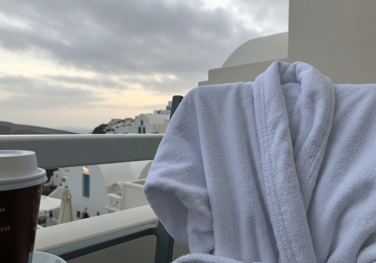 A slightly overcast Santorini morning. A close-up shot from a balcony, focusing on a simple white hotel bathrobe draped over a chair, with a partially visible instant coffee cup and a glimpse of soft dawn light on white buildings in the background. The focus is on the texture and quiet atmosphere, as if taken with a smartphone held casually. Authentic, imperfect lighting