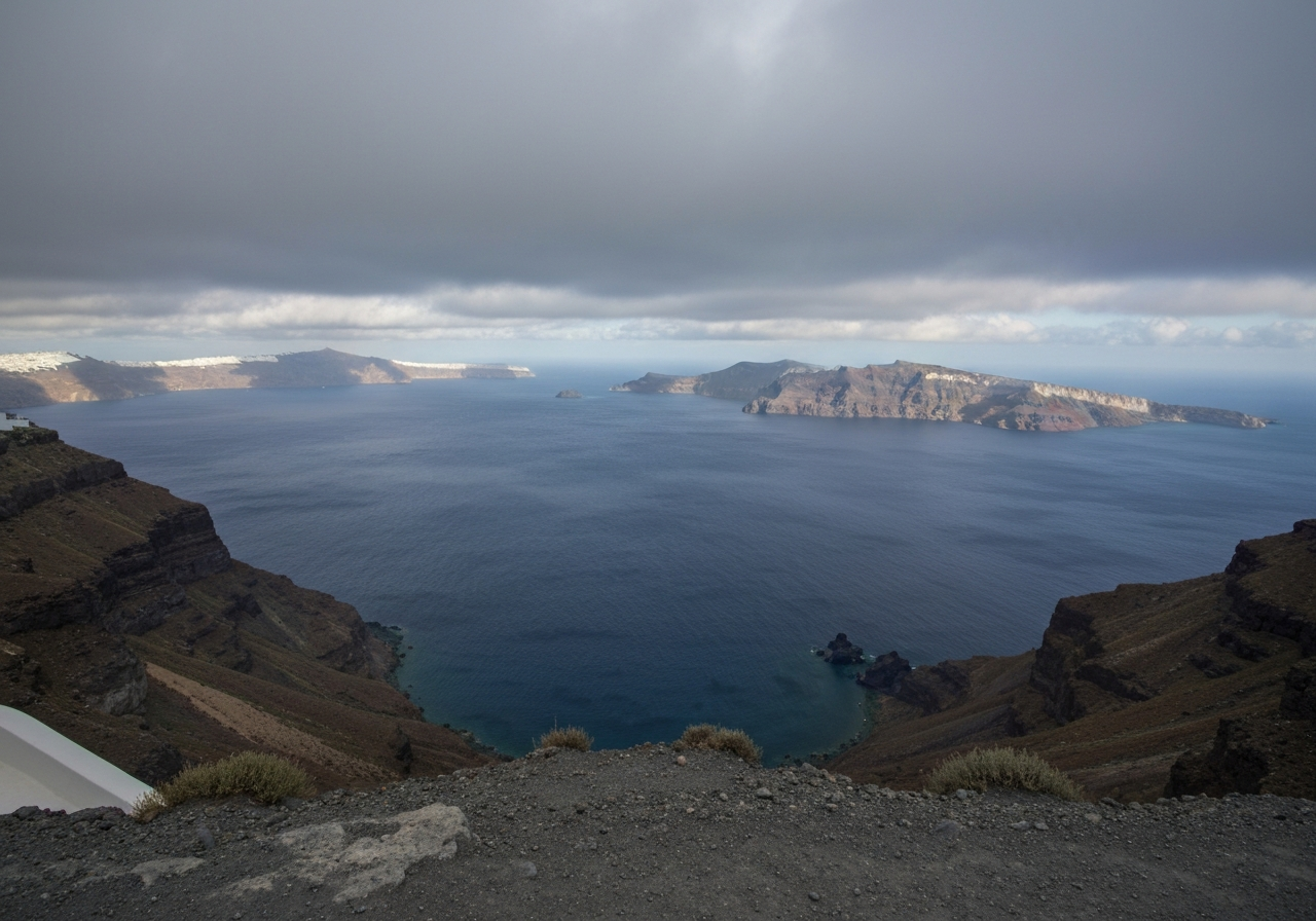 A wide shot from a caldera viewpoint in Santorini on a cloudy morning. The vast caldera stretches out, with dramatic shadows cast across the water and distant islands by the thick cloud cover. The composition is slightly off-center, capturing the raw, powerful landscape rather than a perfect postcard view. The image should feel like a spontaneous capture of the immense geological scale, with a hint of the volcanic rock