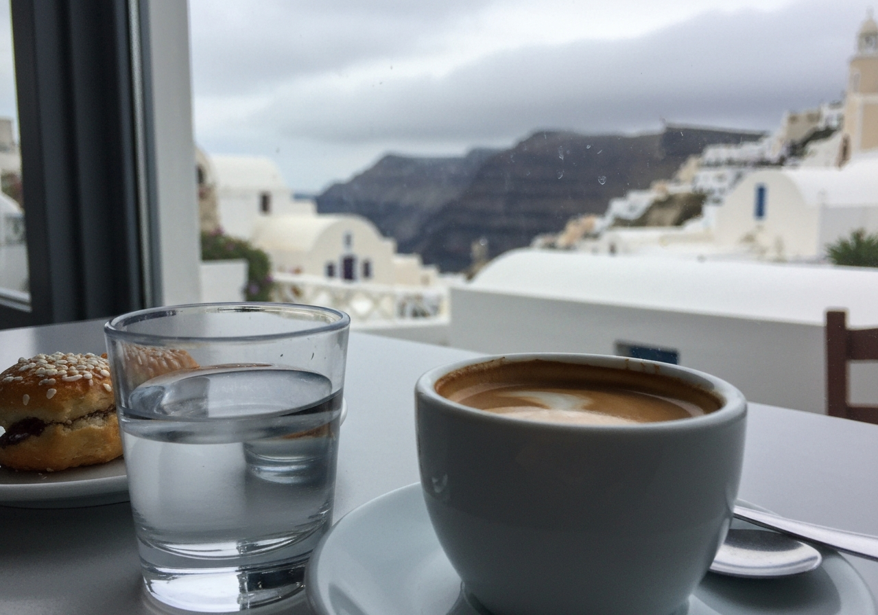 A close-up, slightly shaky smartphone photo taken from a cafe table in Fira, Santorini. The foreground shows a half-finished Greek coffee with a small glass of water and a sweet pastry. In the blurred background, through a slightly smudged cafe window, the cloudy caldera and white-washed buildings are visible. The lighting is soft and diffused, indicative of a cloudy morning. Shot with a modern smartphone, slight lens flare