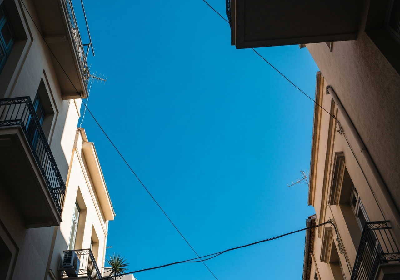 A worm's-eye view looking up at the bright blue Athenian sky from a narrow street. The shot captures the tops of buildings, a few power lines, and perhaps a glimpse of a balcony with a potted plant. The light is strong, casting subtle shadows, and the composition feels spontaneous, as if the photographer quickly looked up to capture the feeling of a sunny departure day