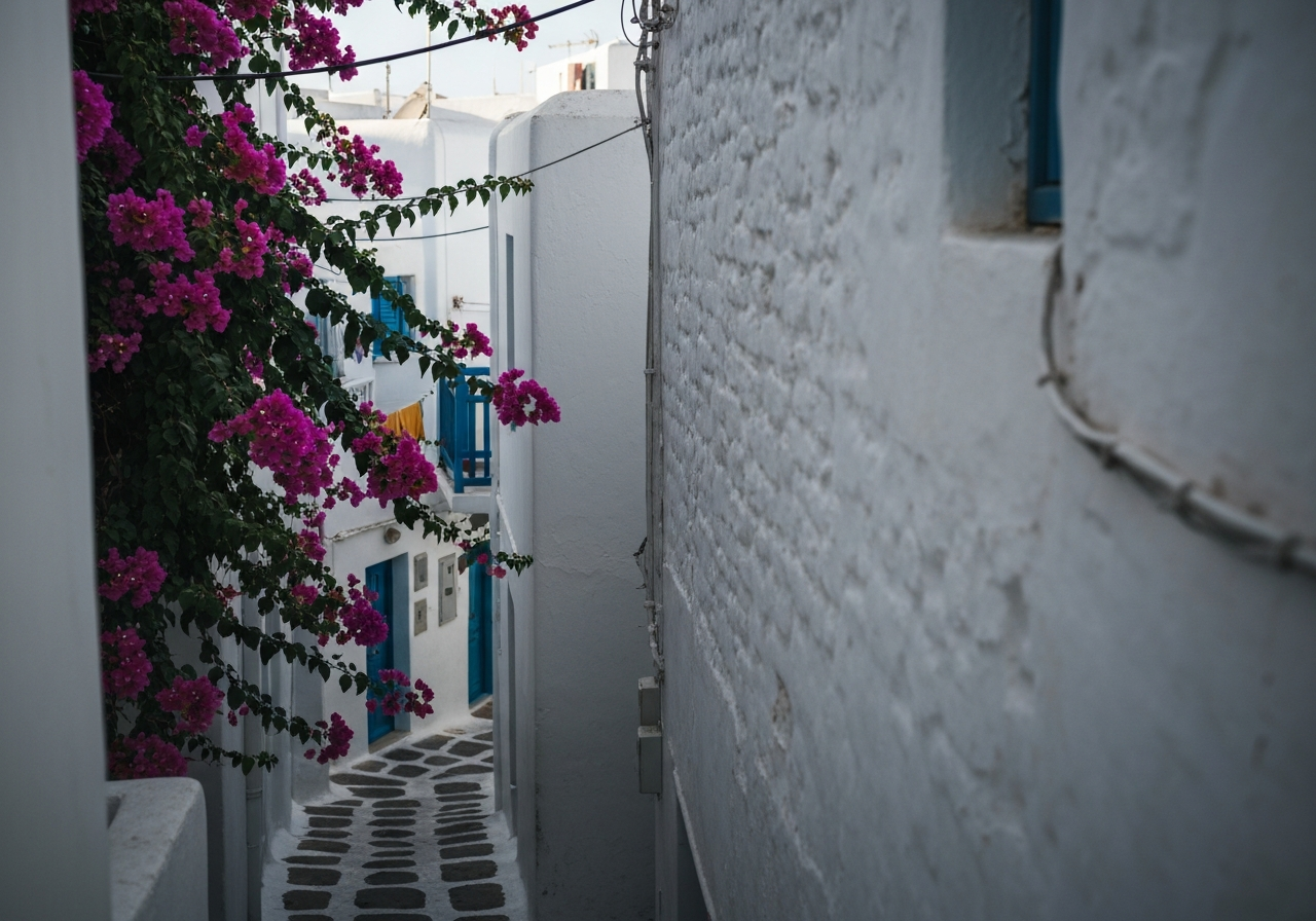 A candid shot taken from a slightly elevated perspective, showing a glimpse of a narrow, winding white alleyway in Mykonos Town. The focus is on the texture of the whitewashed walls and the vibrant bougainvillea cascading down. A hint of a resident's laundry hanging from a balcony or a glimpse of a colorful door adds an element of everyday life. The light is dappled, suggesting late morning or early afternoon sun