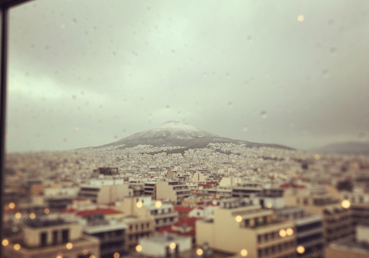 A slightly overcast sky over Athens, with a faint dusting of snow visible on the distant Mount Parnitha, as seen from a hotel window. The image should have a slightly grainy, smartphone-like quality, capturing the unexpectedness of May snowfall