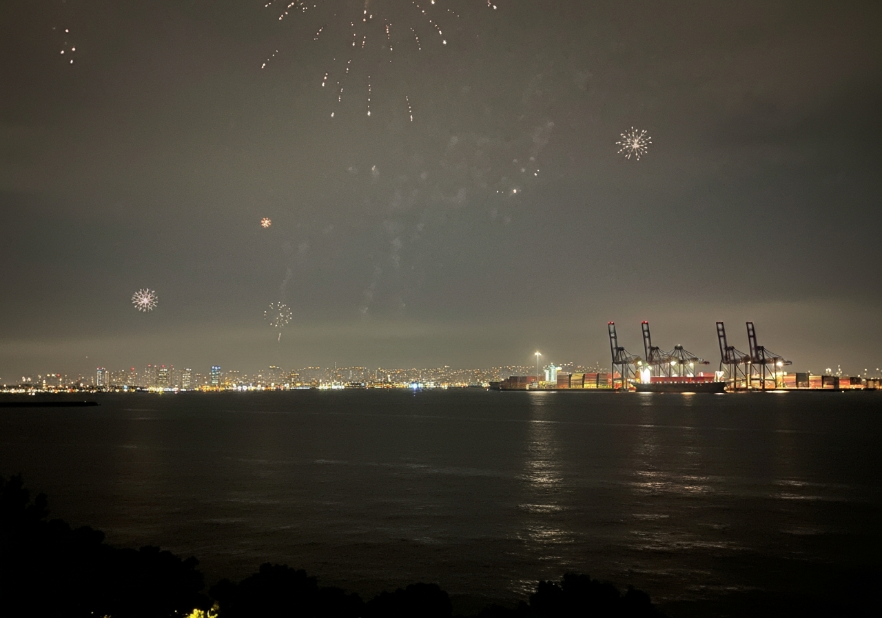 A candid smartphone photo looking across a wide, slightly hazy expanse of water towards a distant, sprawling cityscape at night. The lights of container cranes and ships are visible, creating a utilitarian and industrial feel. The sky above is mostly dark, with a subtle, diffused glow from scattered fireworks that are largely obscured or out of frame. The composition should feel unposed, as if snapped quickly to capture the atmospheric contrast between the quiet foreground and the active port
