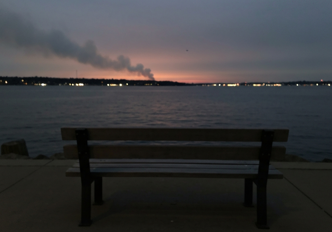 A slightly blurry, handheld smartphone photo taken at dusk of a lone wooden bench facing a dark body of water. Faint wisps of smoke are visible in the distance against a darkening sky, with the subtle glow of distant city lights reflecting on the water. The overall mood is quiet and contemplative, with a focus on the emptiness of the bench and the vastness of the bay