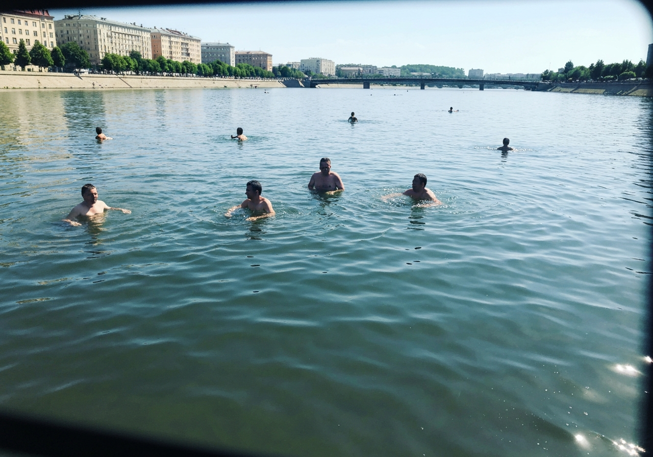 A wide, slightly shaky shot from a moving perspective, showing people swimming and enjoying themselves in a river on a hot day. The water is a murky blue-green, and the sunlight reflects off the surface. In the background, city buildings are visible. The image feels unposed and captures the simple joy of people seeking relief from the heat. Shot on a smartphone, with a natural, slightly imperfect framing