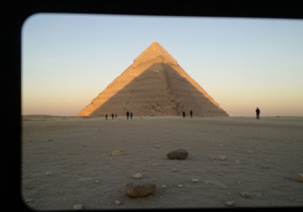 A slightly blurry, handheld shot from a smartphone, taken just after sunrise at the Giza plateau. The golden light is just beginning to hit the top of the Great Pyramid, casting long shadows. A few other early visitors are small figures in the distance, adding a sense of scale. The foreground is a bit dusty, with a few scattered pebbles. Shot with a slightly wide-angle lens