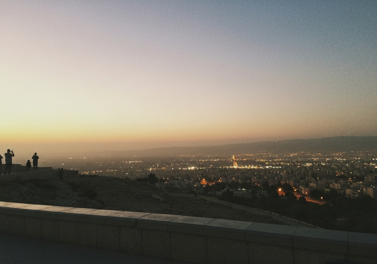 A slightly grainy, low-light smartphone photo taken at a city viewpoint overlooking Amman at dusk. The horizon is a soft blend of fading golden light and the emerging artificial glow of the city. A few indistinct figures might be visible in the distance, also looking out at the view. The image conveys a sense of urban vastness and the subtle transition from day to night, with a hint of atmospheric haze