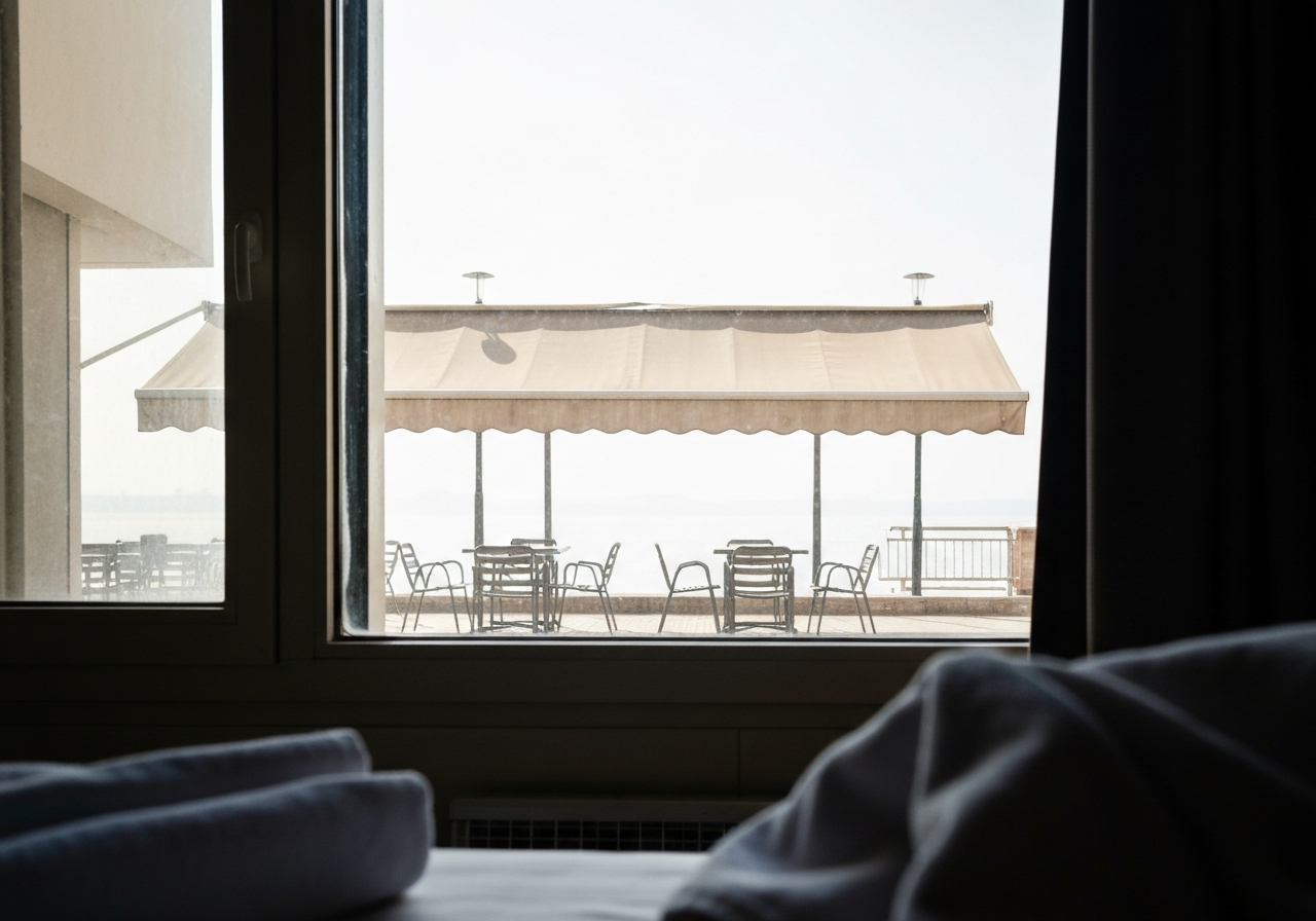 A view through a slightly smudged hotel window, looking out at a deserted street cafe. A few empty tables and chairs sit under a faded awning. The sunlight outside is a bright, almost blinding white. In the foreground, a corner of a hotel bedspread or a discarded cold towel is visible, hinting at someone seeking refuge inside. The image should convey a sense of stillness and the oppressive mid-day heat