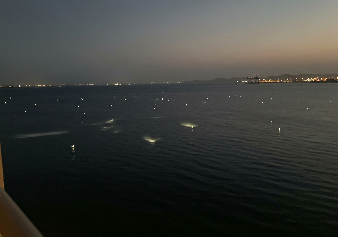 A slightly blurry, handheld smartphone photo taken at dusk from a hotel balcony overlooking the Red Sea in Aqaba, Jordan. The water is dark, reflecting scattered, wavering lights from a distant port. A gentle breeze is visible in the slight ripple on the water's surface. The mood is peaceful and contemplative. Shot with a modern smartphone, natural lighting, slight motion blur