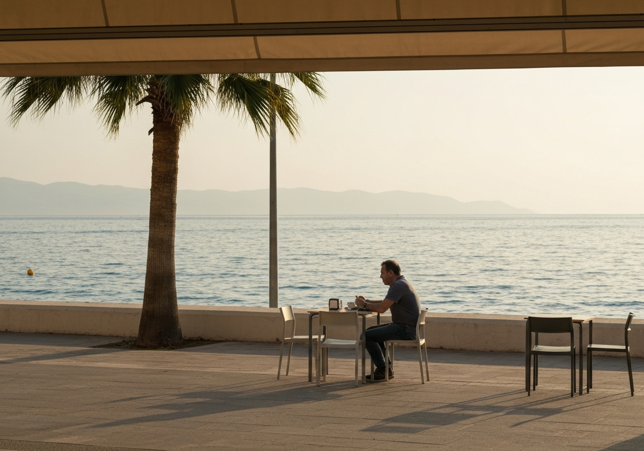 A wide shot from a slightly elevated perspective, capturing a person sitting at a simple outdoor cafe table on a waterfront promenade. The sea is visible in the background, bathed in the warm, hazy light of late afternoon. A few palm trees are in the foreground, and distant mountains are visible across the water. The image should feel relaxed and observational, as if captured spontaneously