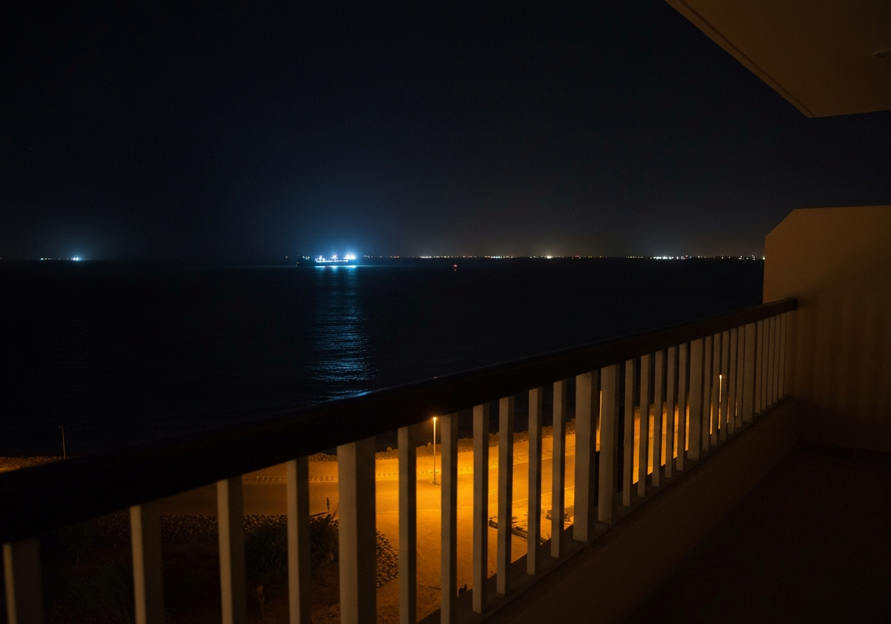 A wide-angle smartphone shot of a dimly lit balcony railing at night. Beyond it, the dark expanse of the Red Sea is visible, with scattered, diffused lights from a port or a moving vessel. The image should feel intimate and solitary, capturing a moment of quiet reflection