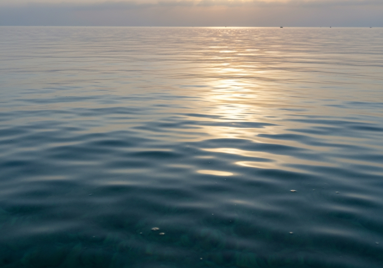A close-up, handheld shot of the calm Red Sea surface in the early morning light. The water appears unnaturally smooth, almost like polished glass, reflecting the soft, developing sky. The light catches the water in a way that suggests solidity, with subtle imperfections like a slight ripple or a stray reflection from a distant boat. It feels like a moment of quiet observation on a smartphone