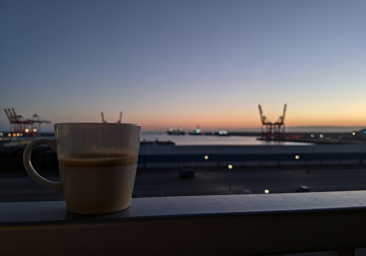 A slightly out-of-focus, candid shot taken from a balcony at dawn. The foreground shows a half-empty, cold cup of coffee on a simple railing. In the background, the pre-dawn light is subtly transitioning from deep indigo to pale orange, illuminating a quiet port with a few distant ships and cranes. The overall mood is contemplative and still, as if captured by a smartphone with a slight shake
