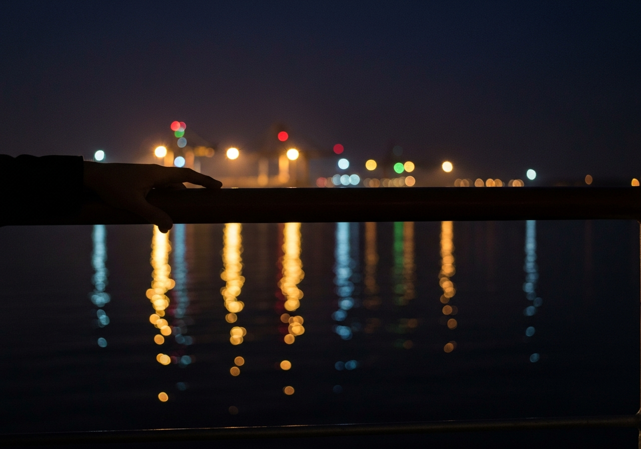 A slightly shaky, low-light smartphone photo taken from a balcony at night. In the foreground, a hand rests on the railing, silhouetted against the faint glow of distant port lights. The lights create abstract, broken reflections on dark water. The overall mood is contemplative and quiet