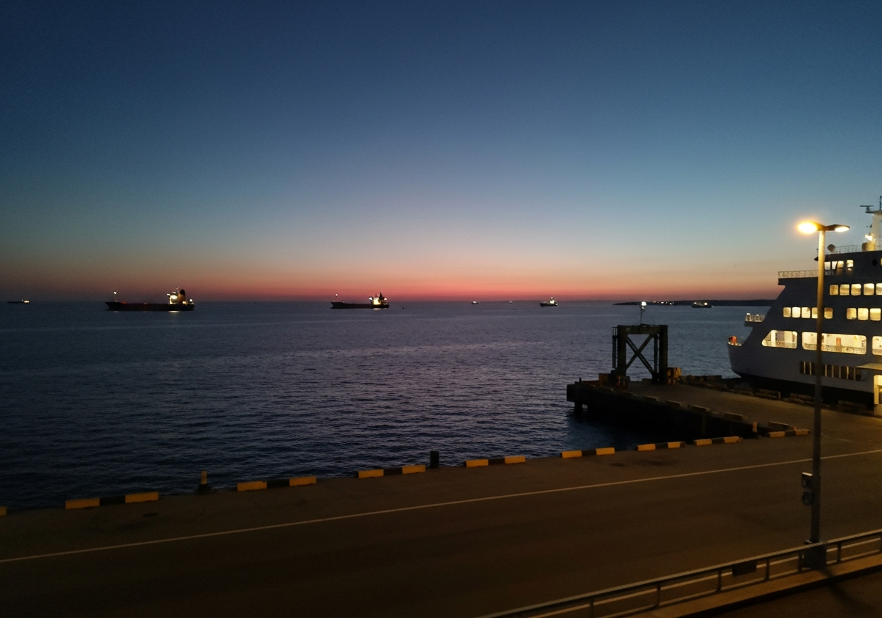 A slightly blurry, handheld smartphone photo taken at dawn, looking out over a quiet ferry terminal. The water is dark with the first hints of orange and pink on the horizon. A few large cargo ships are visible in the distance, their lights twinkling. The scene feels still and expectant, capturing the quiet before the day's activity begins. Shot with a modern smartphone, natural light